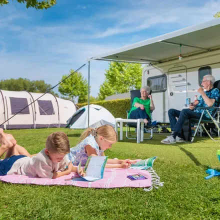 Twee kinderen kleuren op een kleed in het gras, met erachter grootouders bij hun caravan en een tent op de kampeerplaats